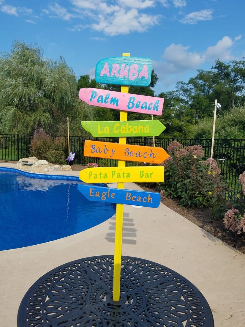 A hand-painted Aruba directional sign standing beside a backyard pool under a blue sky