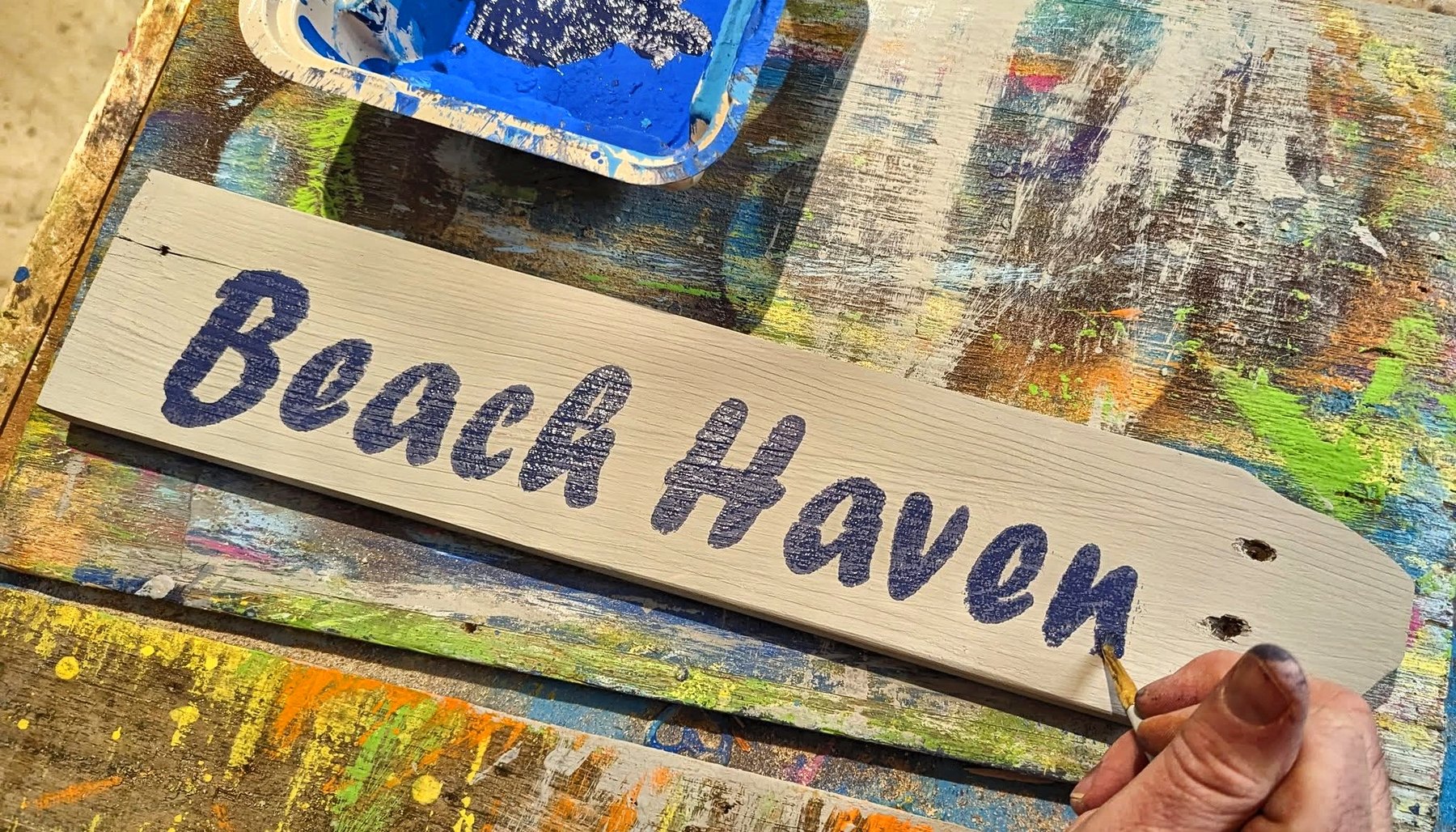 A hand painting the words Beach Haven onto a reclaimed wood sign on a paint-covered workbench